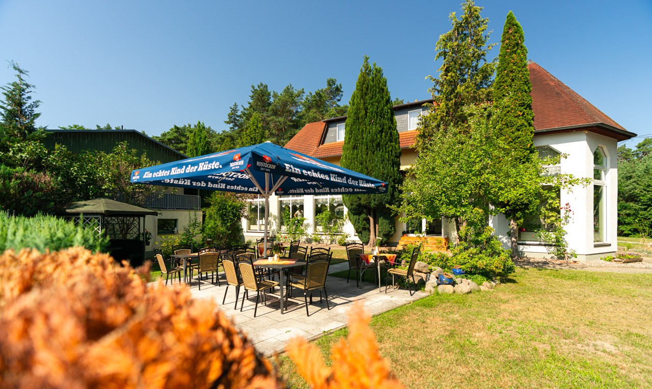 Biergarten auf einer Terasse mit Sonnenschirm, dahinter das Gebäude des Natur- und Waldhotel Johannesruh umgeben von Grün.