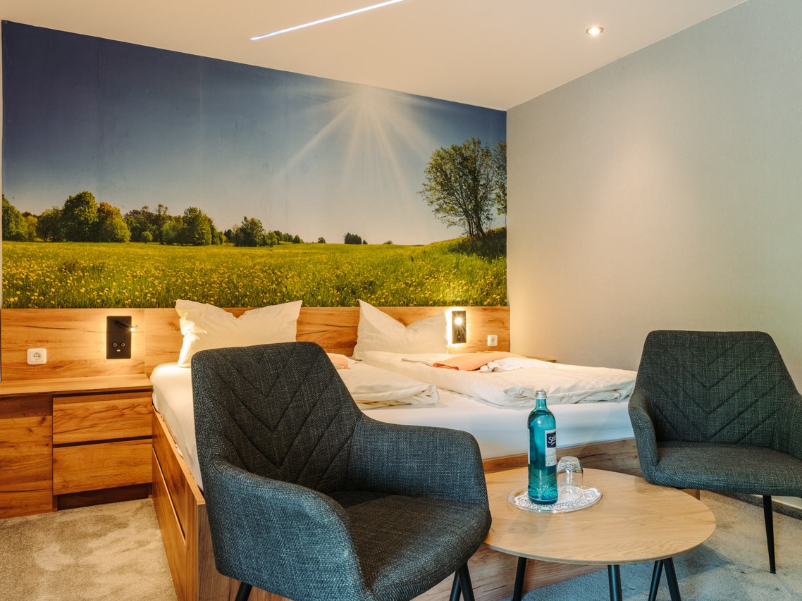 Doppelbett mit zwei Nachttischen in Holzoptik. An der Wand am Kopfende des Bettes ein idyllisches Bild von einer blühenden Wiese am Waldrand mit blauem Himmel und Sonnenschein. Am Fußende des Bettes ein kleiner runder Tisch mit zwei Sesseln. Teppichboden in Beige.