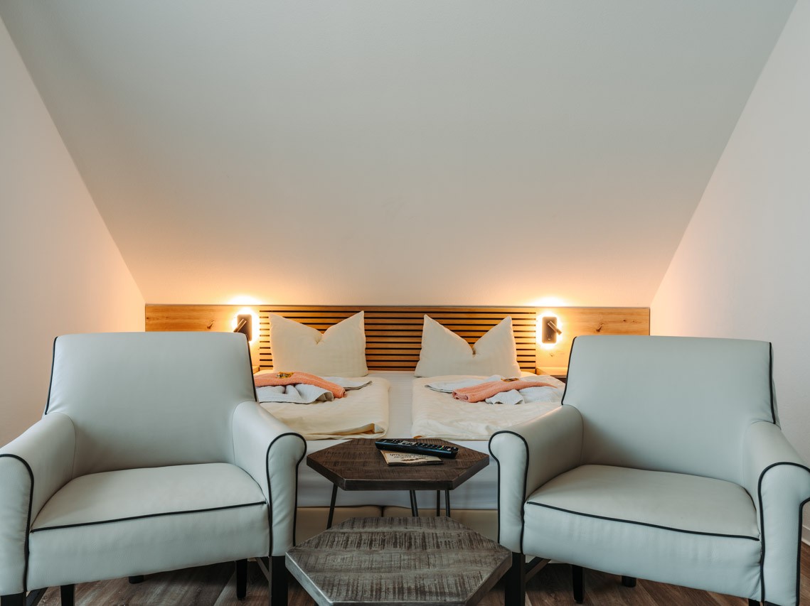 Zimmer mit Dachschräge. Unter der Schräge ein großes Doppelbett mit integrierten Nachttischen. Am Fußende des Bettes zwei Sessel mit einem Beistelltisch.